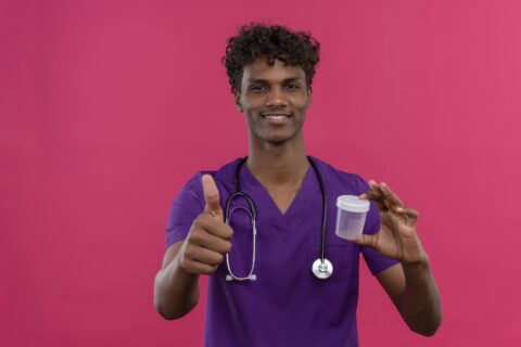 a happy young handsome dark-skinned doctor with curly hair wearing violet uniform with stethoscope showing thumbs up while holding plastic specimen jar on a pink background