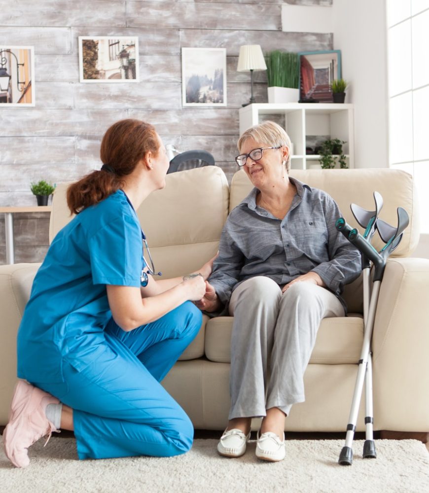 Happy old woman in a nursing home sitting on couch talking with her caretaker. Retired woman with crutches.